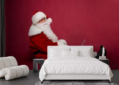 Happy Santa Clause working on laptop while sitting on red background, Christmas and New year concept Wall mural