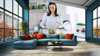 Happy smiling cute woman is preparing a fresh healthy vegan salad with many vegetables in the kitchen at home and trying a new recipe Wall mural