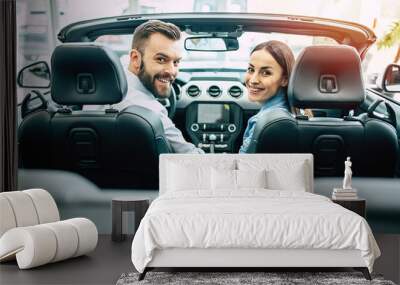 Happy young couple chooses and buying a new car in the dealership and looking on camera. Wall mural