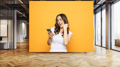 Hell yes! Screaming from good news girl with nice hairstyle and perfect skin in front of spicy-orange color is holding her phone and smiling sincerely. Wall mural