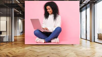 Hit the ground running. Magnificent African American lady in a snow-white sweater is sitting cross-legged with her laptop, smiling happily while looking at the screen. Wall mural