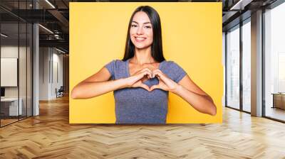 Love, heart shape, peace. Sign, gesture or symbols. Portrait of cute attractive smiling brunette woman, girl shows heart sign with help of her hands on yellow background in casual wear Wall mural