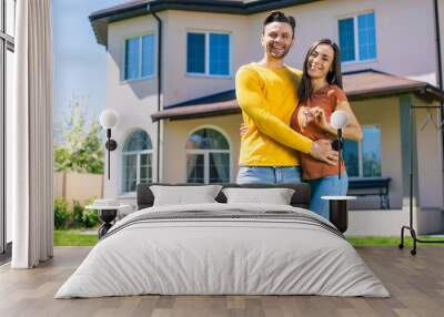 Modern beautiful excited young couple in love standing in front of the new big house with the keys in hands and hugging while they celebrating this buying Wall mural