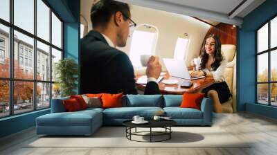 Movers and shakers. A lovely woman in formal attire is typing something on her laptop and drinking coffee while sitting near her male colleague in a window seat in a business class. Wall mural