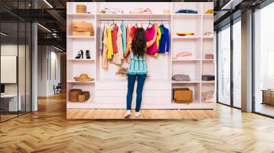 My new clothes. A fit girl in summer clothes is standing with her back to the camera, trying to choose an outfit to wear from an abundance of clothes in her compartment wardrobe. Wall mural