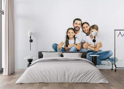 Relationship concept. Beautiful and happy smiling young family in white T-shirts are hugging and have a fun time together while sitting on the floor and looking on camera. Wall mural