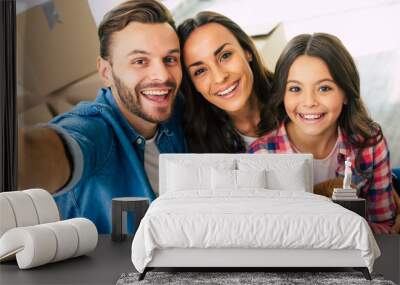 Smile, my dear! Close up photo, joyful family is taking selfie together in their new house in front of a pile of cardboard boxes. Wall mural