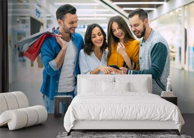 Stylish happy excited friends with shopping bags are looking together on the smart phone and chatting about sales in the mall Wall mural