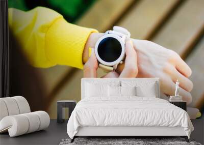 Turning smartwatch. A close-up photo from behind of a girl in a yellow hoodie, with a white modern smartwatch, turning it on with two fingers. Wall mural