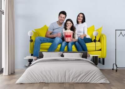 Watching comedies. Happy mother, father and their little cute daughter are sitting together on a yellow couch and laughing while watching comedies. Wall mural