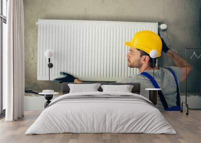 Young handsome professional plumber in modern uniform is installing heat radiator in a new building under window Wall mural