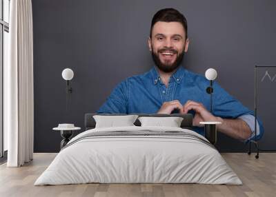 Young handsome smiling bearded man in denim shirt shows hands in the shape of hearts and looks on the camera Wall mural