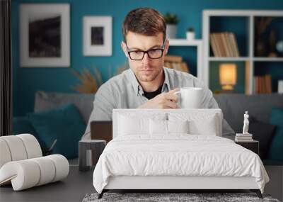 Front view of involved young man browsing laptop and holding cup sitting at table in living room Wall mural