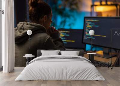 Pensive mature woman in eyeglasses sitting ast home office with two modern computers while programming. Dark atmosphere during evening time. Wall mural