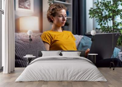 Young curly woman wear glasses using laptop to communicate with customer, or write email to office from home seating on couch in light room Wall mural