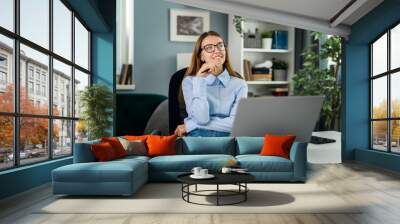 Young thoughtful woman wearing eyeglasses dreaming at desk with laptop, motivated idea concept, smiling female working in cozy light room Wall mural