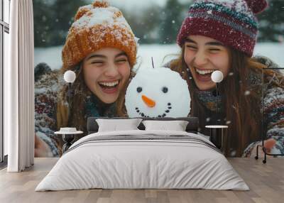 Two young girls laugh and pose with a snowman they have built together, surrounded by a beautiful winter wonderland with falling snowflakes. Wall mural