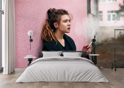 Portrait of a hipster teen girl smoking in a warm black jacket leaning on the loft brick wall, smokes under stress, wanting to relax, letting out smoke cigarettes. Wall mural