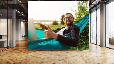 Cheerful bearded smiling young male lying in hammock in park looking to camera satisfied with his ideal dream job Wall mural