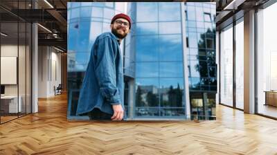 Happy young trendy bearded hipster male dressed in casual denim shirt, red hat and eyeglasses looking away and smiling Wall mural