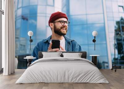 Portrait of happy handsome bearded young male hipster holding smartphone and looking to the side Wall mural