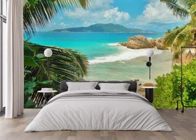 View of turquoise ocean with tropical beach, green vegetation and rocks near the shore. Through green leaves or foliage is an unsharp island visible in the distance. Summer holidays on the Seychelles Wall mural