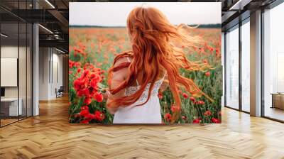 Beautiful young red-haired woman in poppy field with flying hair Wall mural