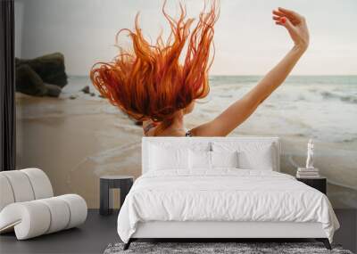 young red-haired woman with flying hair on the ocean coast at sunset, rear view Wall mural