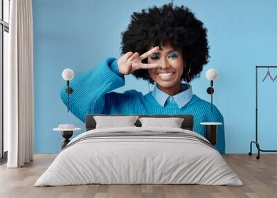 Young, black woman and shows peace sign with blue studio background, trendy style and afro. Smile, happy and relax African American female, lady or girl influencer with casual fashion and cosmetics. Wall mural