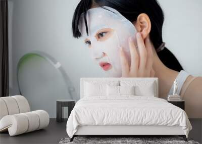 Beautiful young asian woman with sheet facial mask and looking mirror at home, beauty girl applying cosmetic and makeup and touch face with skin care for wrinkle, skincare treatment and moisturizer. Wall mural