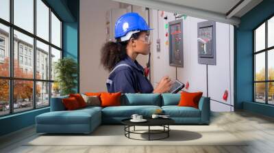 Electrical young asian woman engineer examining maintenance cabinet system electric and using tablet in control room at industrial factory, technician or electrician inspection power distribution. Wall mural