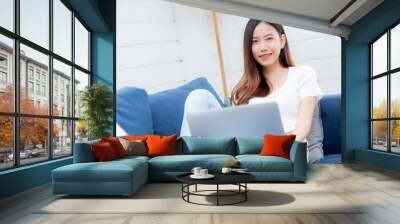 Young asian business woman smile and work from home with laptop computer online to internet on sofa in living room, freelance girl using notebook on couch with comfort, new normal, lifestyle concept. Wall mural