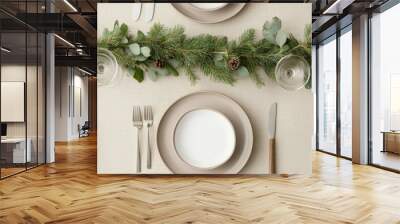 A festive table setting with elegant dinnerware, silverware, and glasses, adorned with a lush green garland centerpiece featuring pine branches and pine cones. Wall mural