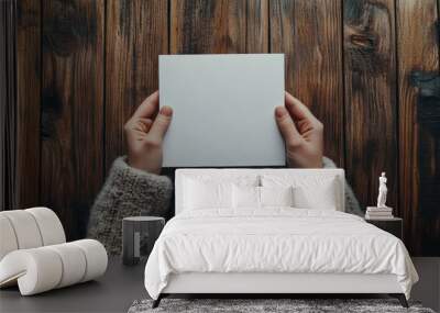 A person holding a blank white square card on a wooden table, dressed in a cozy knit sweater. Wall mural