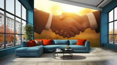 Two people in business attire shake hands warmly against a bright sunset, symbolizing a successful agreement or partnership in a professional context. Wall mural