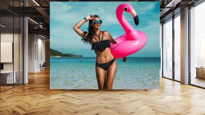 A beautiful sexy amazing young woman on the beach sits on an inflatable pink flamingo and laughs, has a great time, tanned perfect body, long hair, black bikini, fashion accessories, low key photo Wall mural