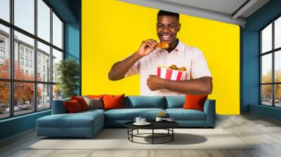 African American young guy eats fast food, chicken leg and a whole bucket in his hands. Emotions of joy and happiness, surprise. Yellow background Wall mural