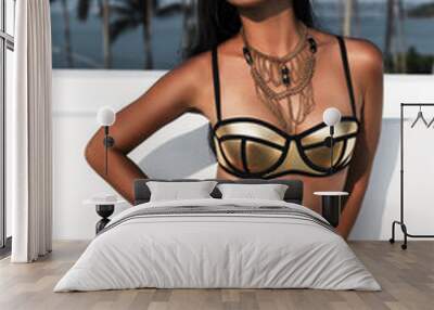 Fashion portrait of a beautiful young sexy woman, brunette. Dressed in a gold swimsuit with gold bracelets and necklaces, tanned skin, an ideal body, against a backdrop of palm trees and a white wall Wall mural