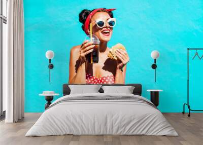 Pretty funny cute girl eating fast food, drinking sodas and eating a hamburger, retro style, blue mint background. cheeky mood, clear hipster glasses, smile, sexy top, fashionable hairstyle with horns Wall mural