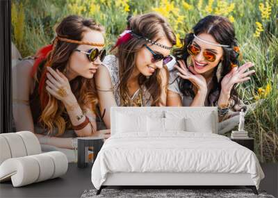 Three cute hippie girl lying on the grass among the wildflowers, best friends having fun and laughing, ukulele, sunglasses, feathers in their hair, bracelets, flash tattoo, indie, Bohemia, boho style Wall mural