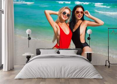 Two beautiful amazing tanned sexy girls, on the beach, laughing smiling having fun, blonde and brunette, long hair, perfect sports figures, fashionable red black swimsuits, sunglasses, tropical island Wall mural