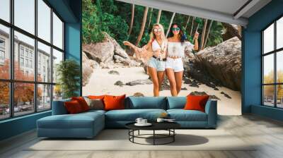 Two cute beautiful girls in Boho style dressed shirts shorts light cape bracelets necklaces, in the hair decorative feathers colored, go embracing the beach against the sea and the rocks Wall mural