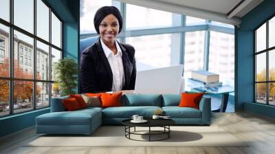 African business woman smiling at camera while she was busy working on her laptop in the business lounge of her modern co working office, with large windows and the cityscape in the background. Wall mural