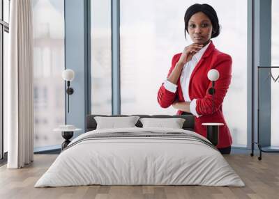 Confident businesswoman looking at the camera with bold body language while wearing a red blazer with large windows behind her in the background. Wall mural