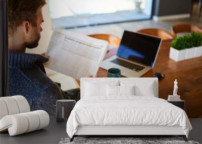 over the shoulder image of an attractive young man holding a newspaper while seated at his beautiful wooden dining table at home, with a hot cup of tea and his laptop ready to work. Wall mural