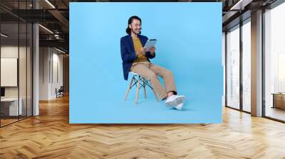 Business man asian happy smiling using a digital tablet while sitting on chair isolated on bright blue background. Wall mural