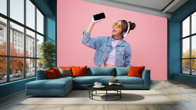 Happy beautiful Asian teen woman in yellow glasses listening music in headphones and dance on pink background. Wall mural