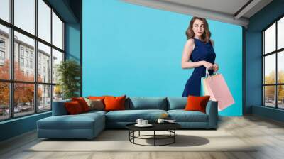 Happy beautiful young woman in blue dress and hand holding shopping bag at copy space isolated over blue background. Wall mural