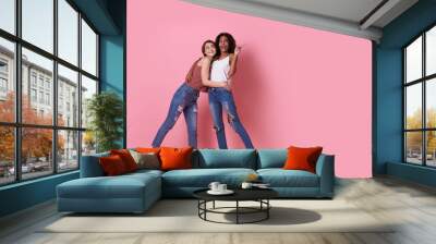 Portrait of two joyful young woman standing with her finger pointing at copy space isolated over pink banner background. Wall mural