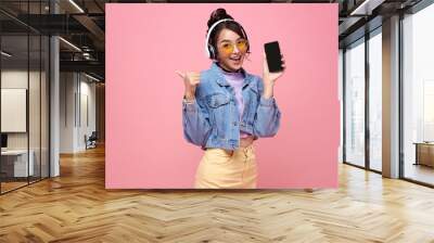 Young Asian teen woman showing smart phone she listening music in headphones isolated on pink background. Wall mural